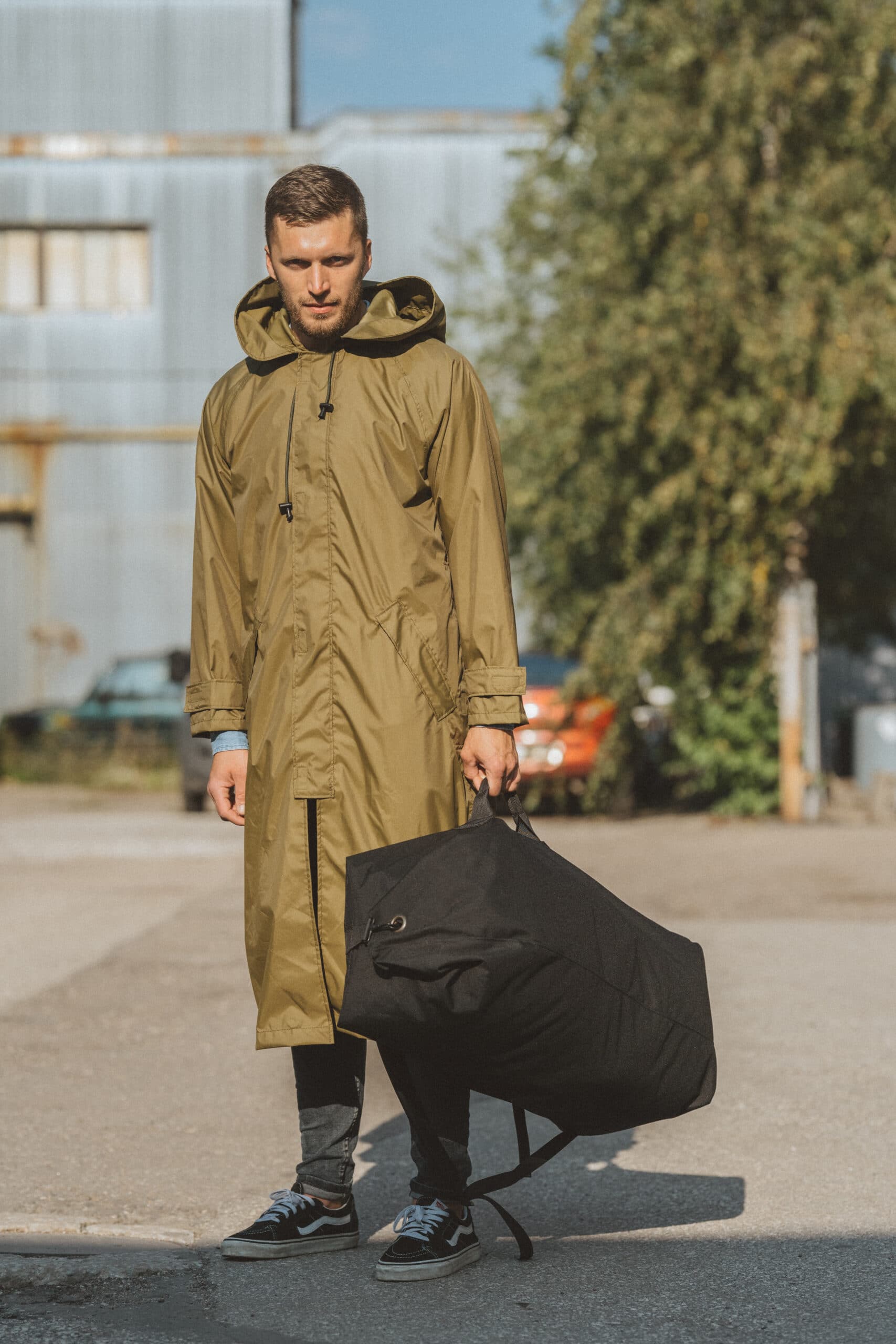 Stylish man holding a black travel backpack while wearing a green raincoat in an outdoor setting
