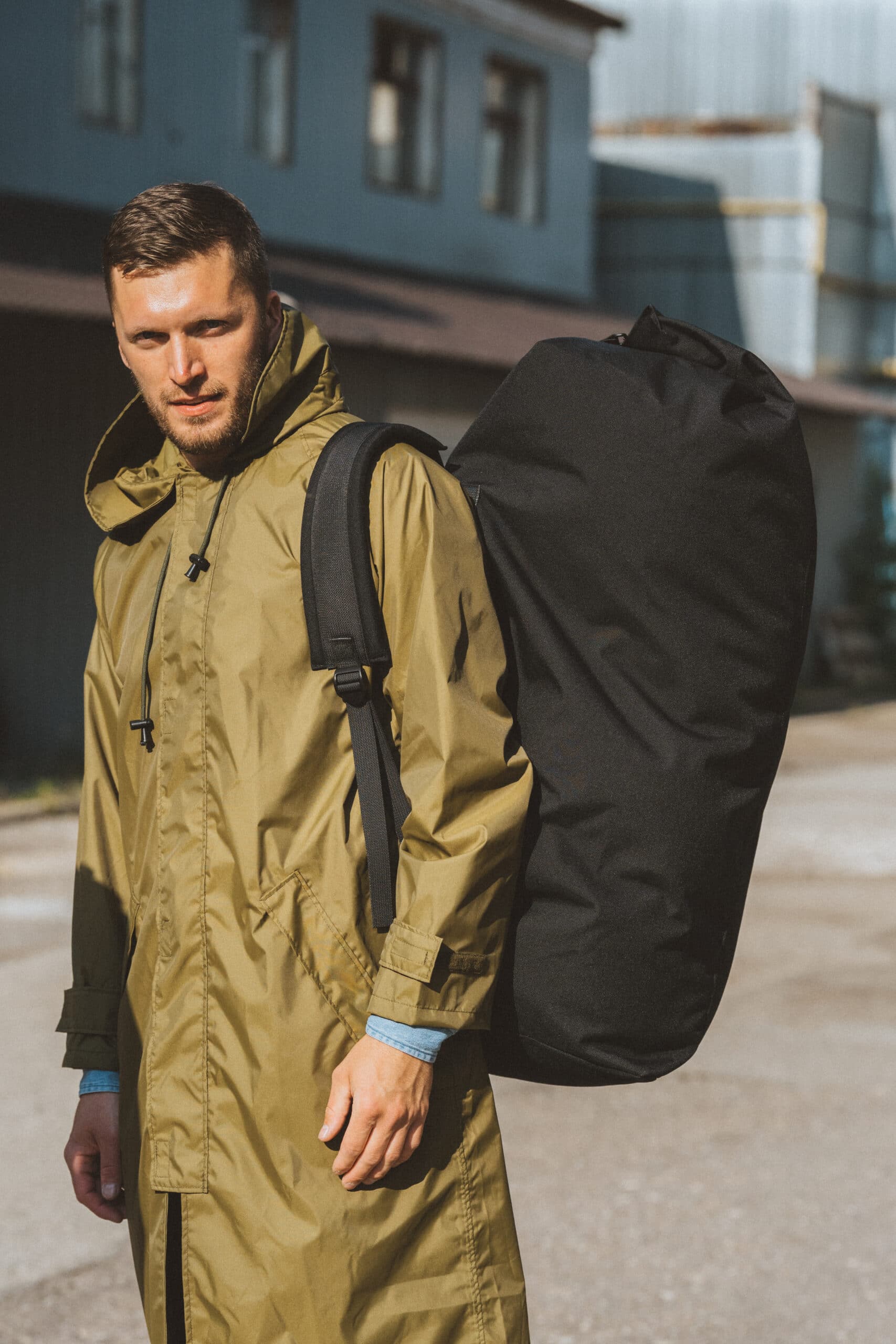 Man wearing an olive green coat with a black travel backpack slung over one shoulder outdoors