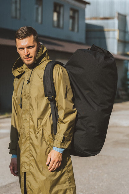 Man wearing an olive green coat with a black travel backpack slung over one shoulder outdoors